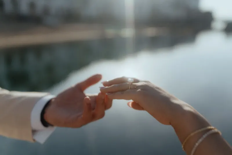 A romantic surprise proposal photoshoot by the water, capturing the moment of slipping on the engagement ring.