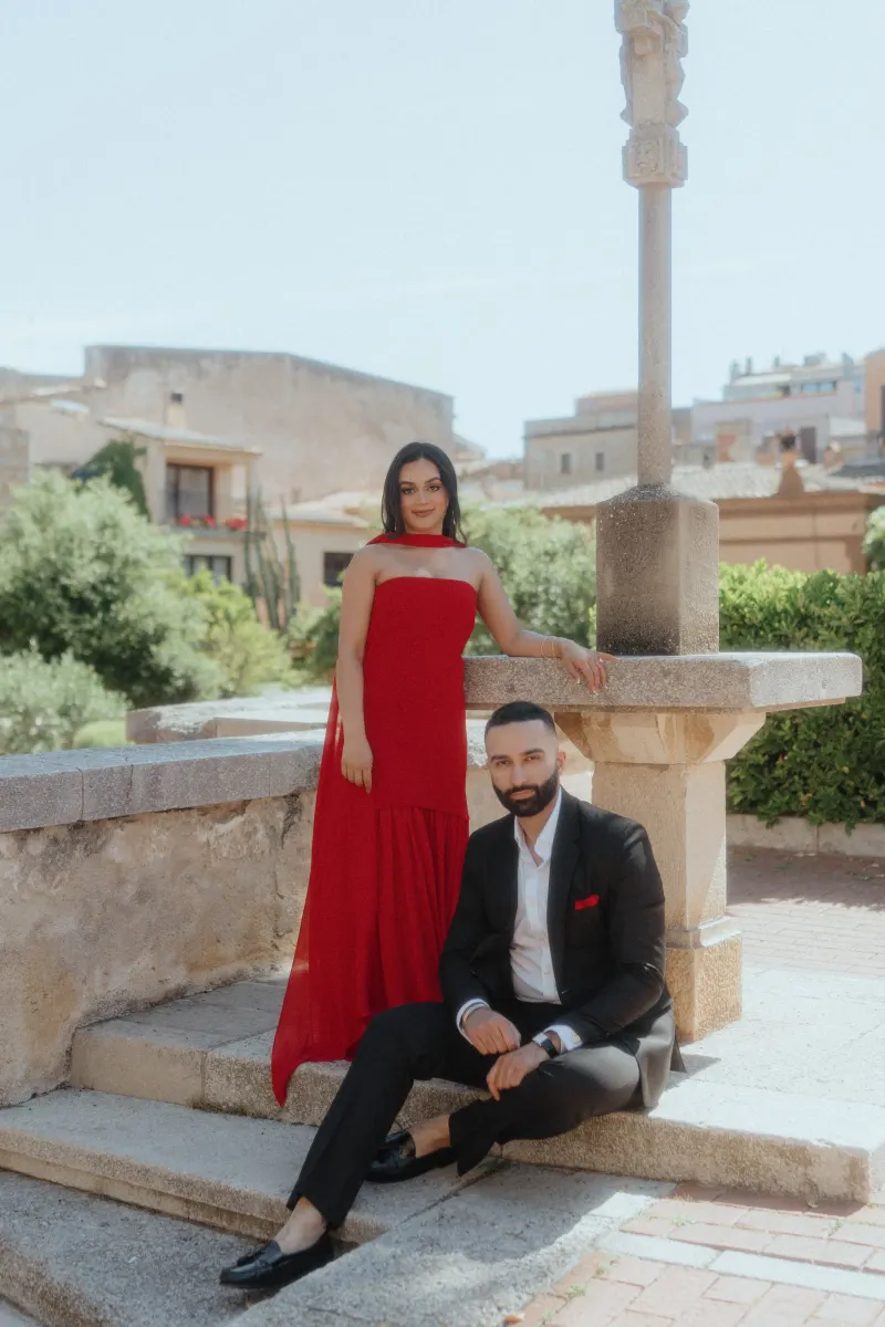 A stylish couple poses by a stone fountain in Begur, dressed elegantly for their surprise proposal photoshoot.