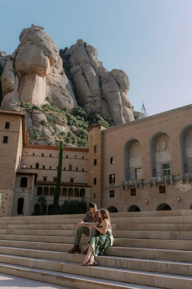 A wide portrait of Tim and Megan seated below the monastery buildings and rocky peaks.