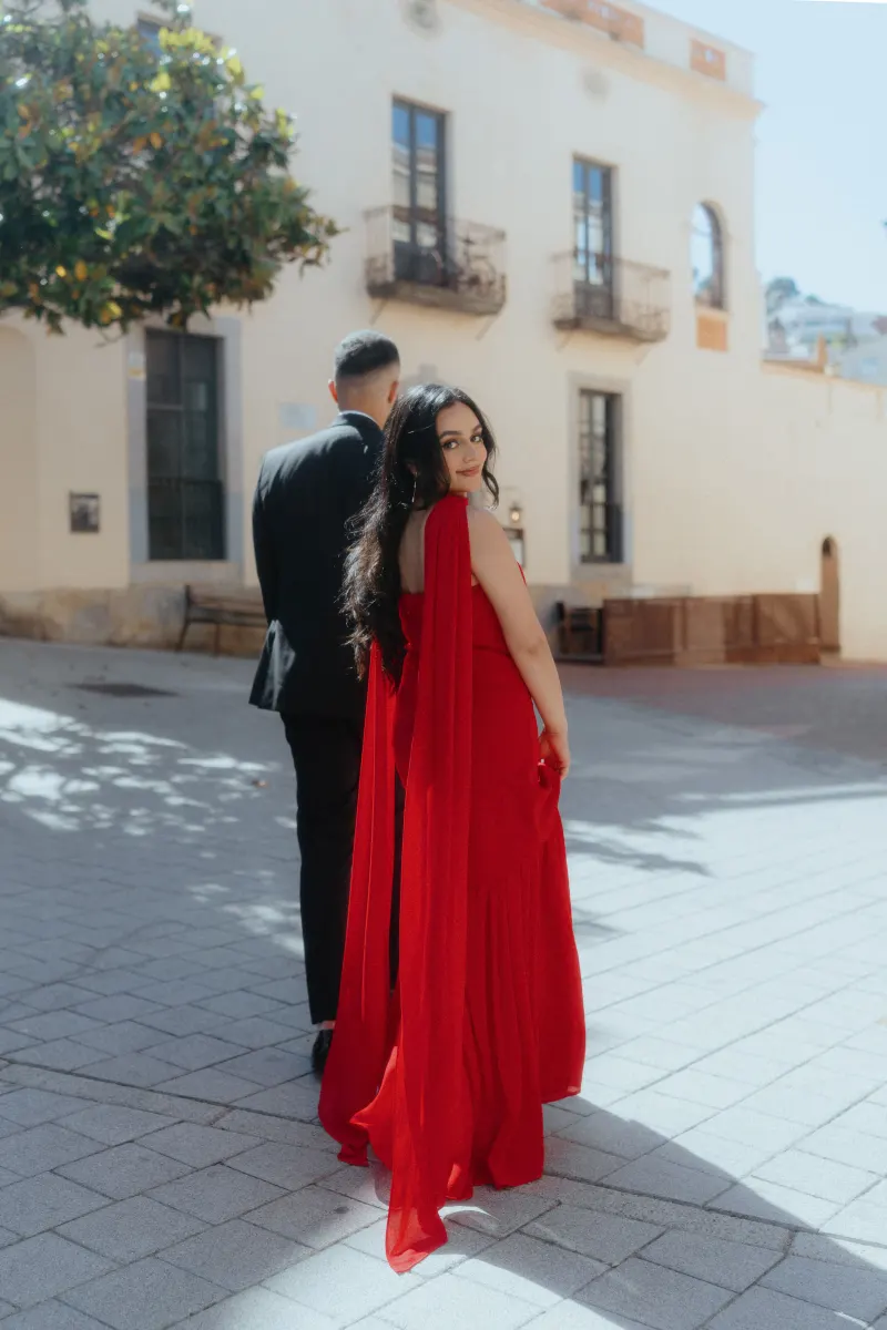 A woman in a flowing red dress glances back, sharing a joyful moment with her partner during a surprise proposal photoshoot.