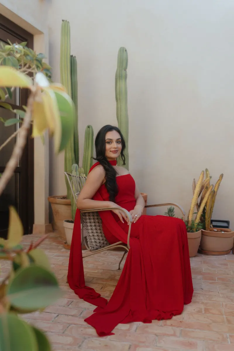 A woman in a flowing red dress sits gracefully on a patio, captured during a surprise proposal photoshoot.