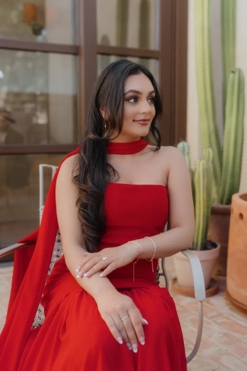 A woman in a red dress sits on a sunlit patio, smiling softly during her surprise proposal photoshoot.