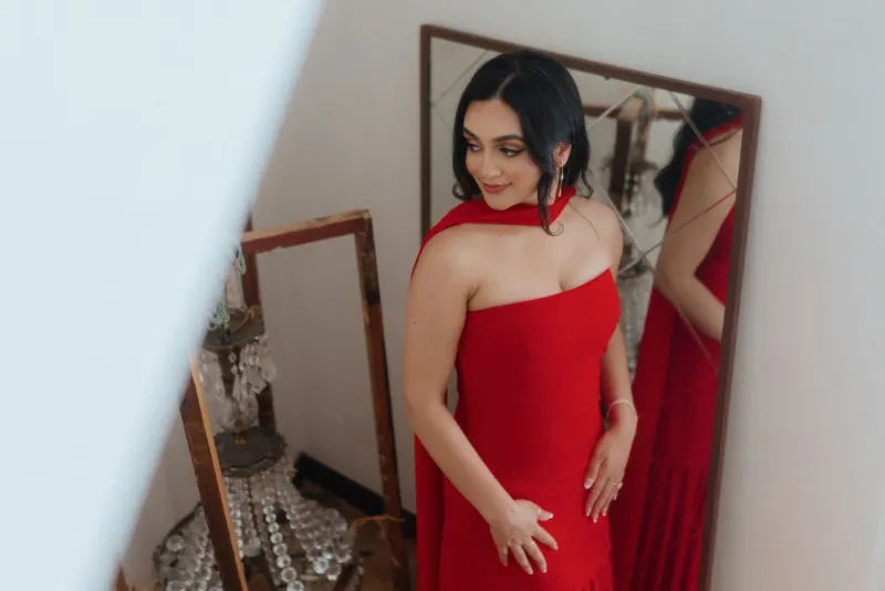 A woman in a red dress smiles softly at her reflection, captured before her surprise proposal photoshoot.