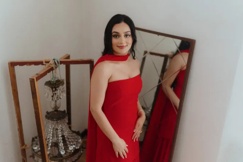 A woman in a red dress smiles softly by a vintage mirror, captured before her surprise proposal photoshoot.