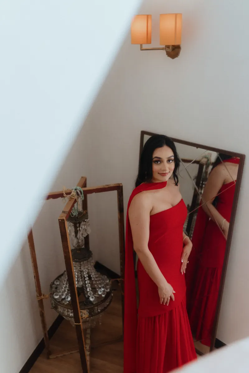 A woman in a red gown stands by a mirror, radiating anticipation before her surprise proposal photoshoot.