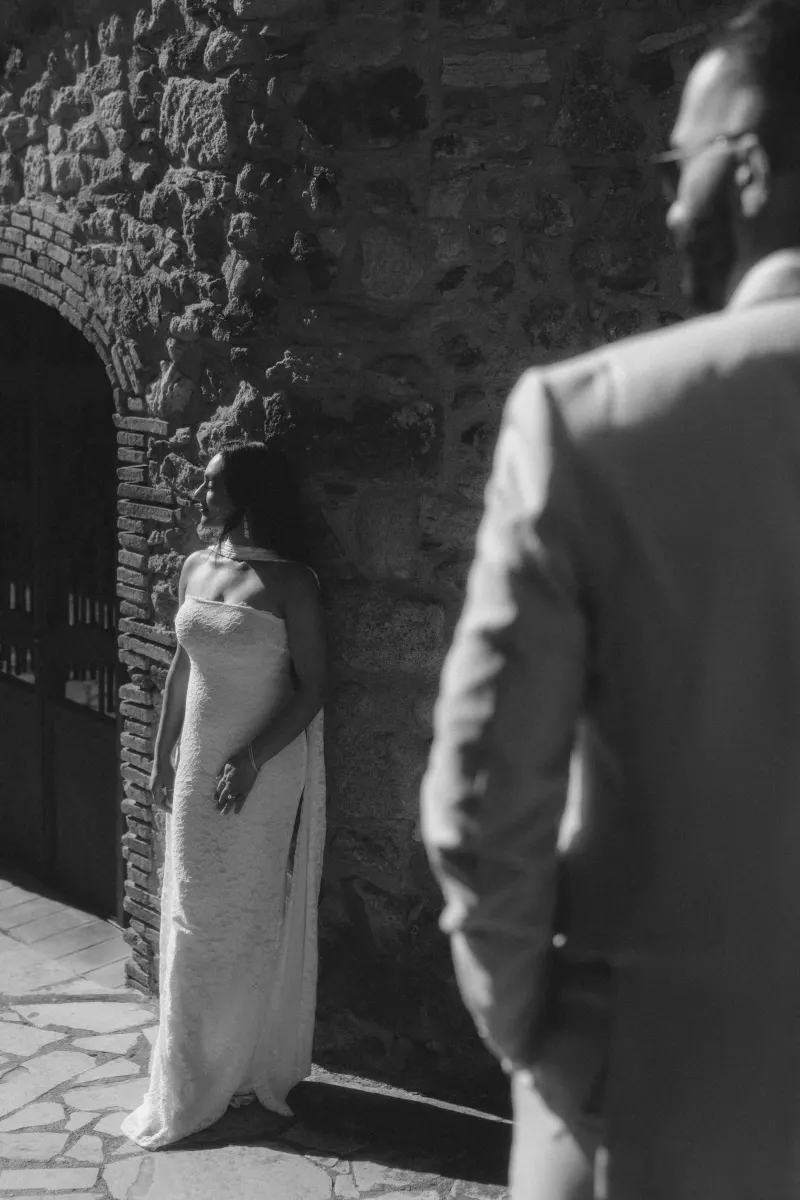 A woman in a white dress smiles against a stone wall as her partner approaches during a surprise proposal photoshoot.