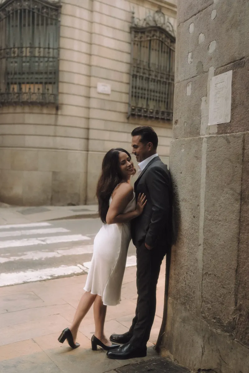 Ashley in a white dress leans toward Edgar on a quiet street corner in the Gothic Quarter.