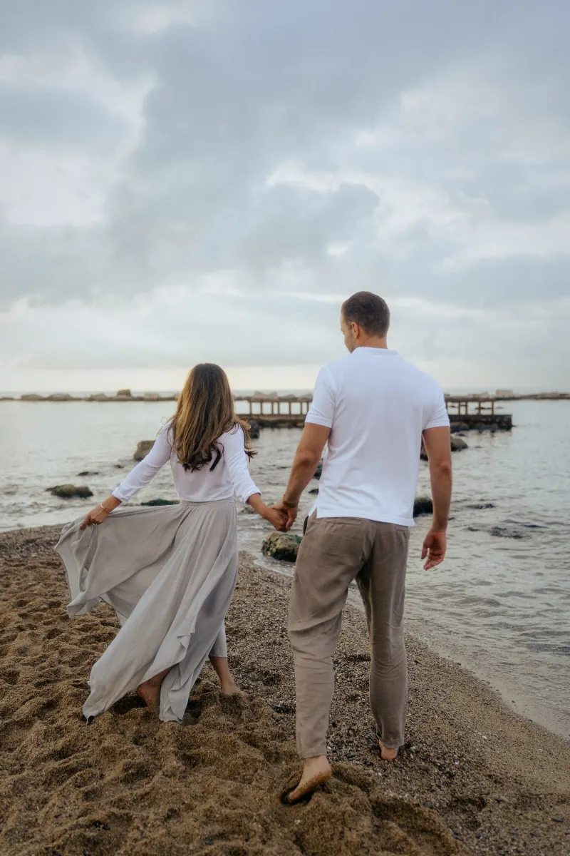 Barefoot couple walking hand in hand along a quiet beach, captured during a couples & engagements photoshoot.