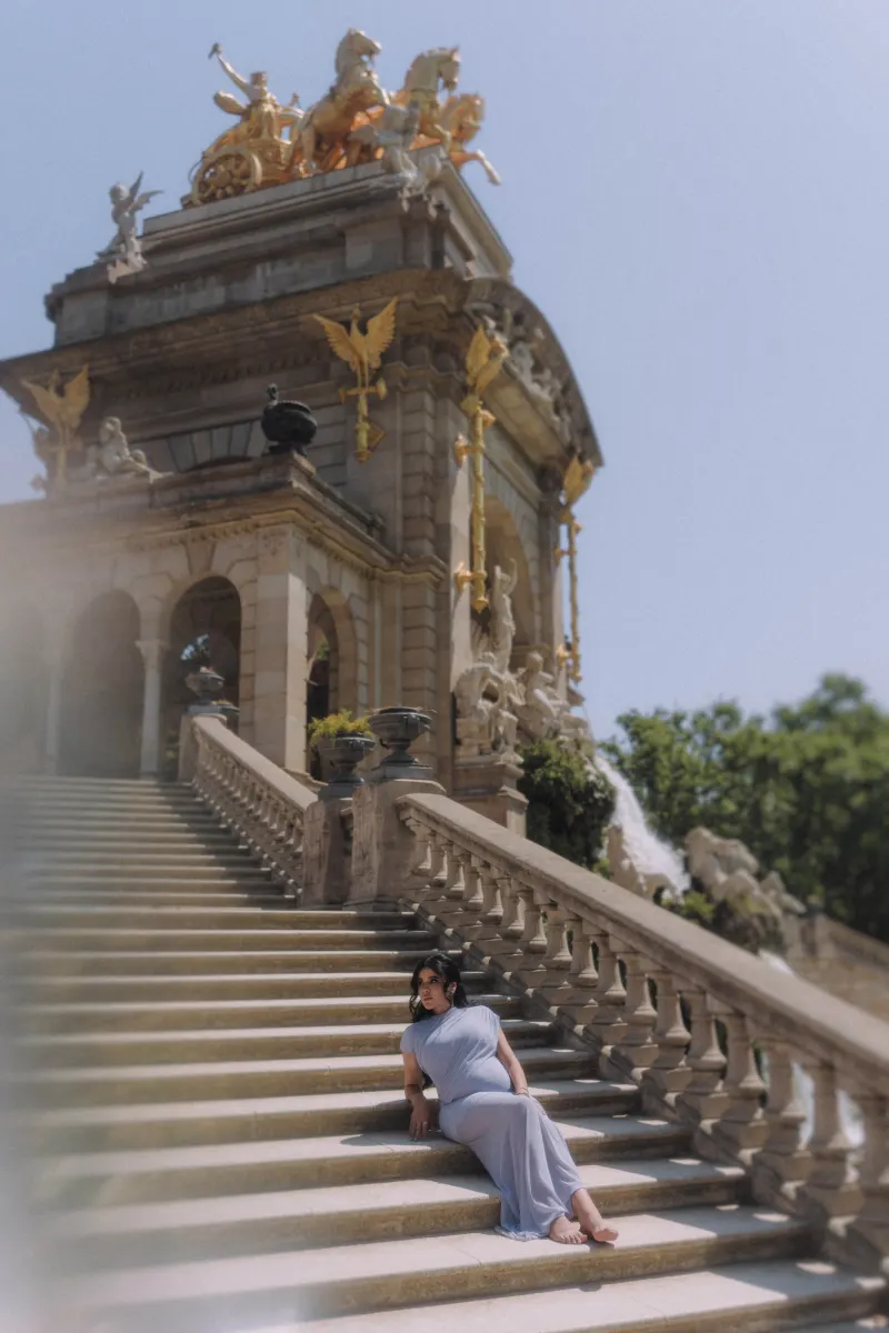 Barefoot pregnant woman in a soft blue gown relaxes on grand stone steps, Vacation Portraits shoot in Barcelona.