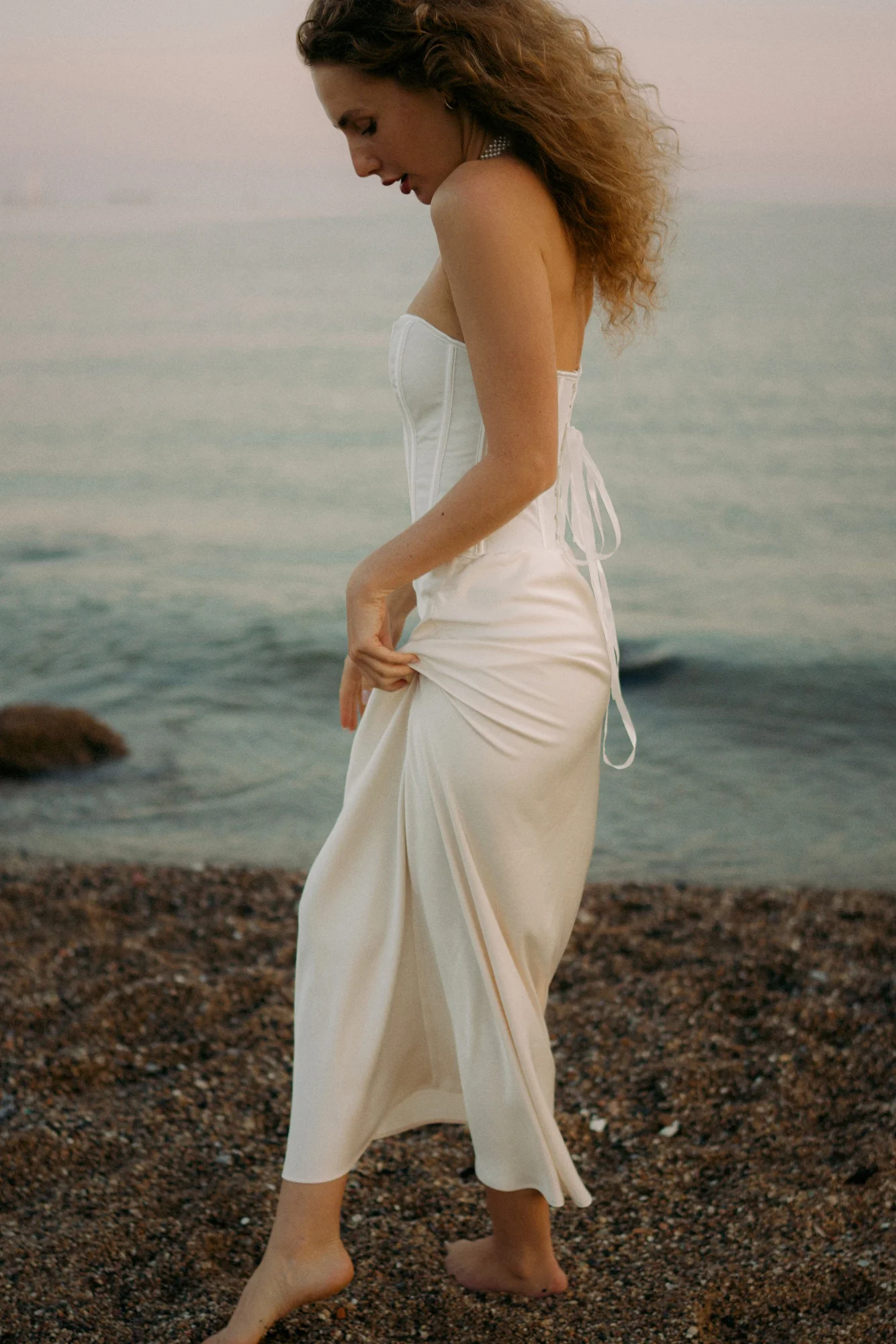 Barefoot woman in a white dress strolls along the shore at sunset during a Vacation Portraits photoshoot.