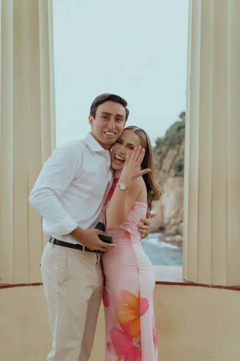 Beaming couple celebrates their engagement at a seaside pavilion, captured during a surprise proposal photoshoot.