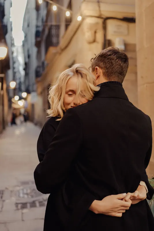 Blonde woman smiles with eyes closed as she hugs her partner on a softly lit Barcelona street during an engagement session.