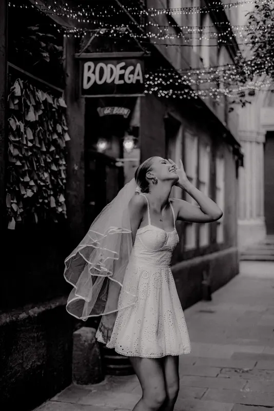 Bride-to-be in a playful white dress laughs under string lights, Vacation Portraits photoshoot in a charming city alley.
