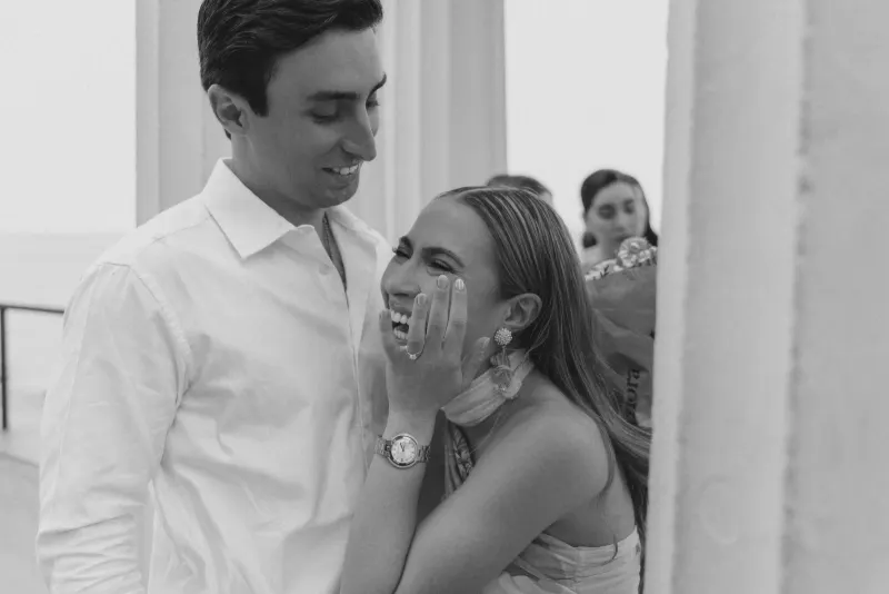 Bride-to-be laughs with joy, showing her ring as her partner smiles beside her during a seaside surprise proposal session.