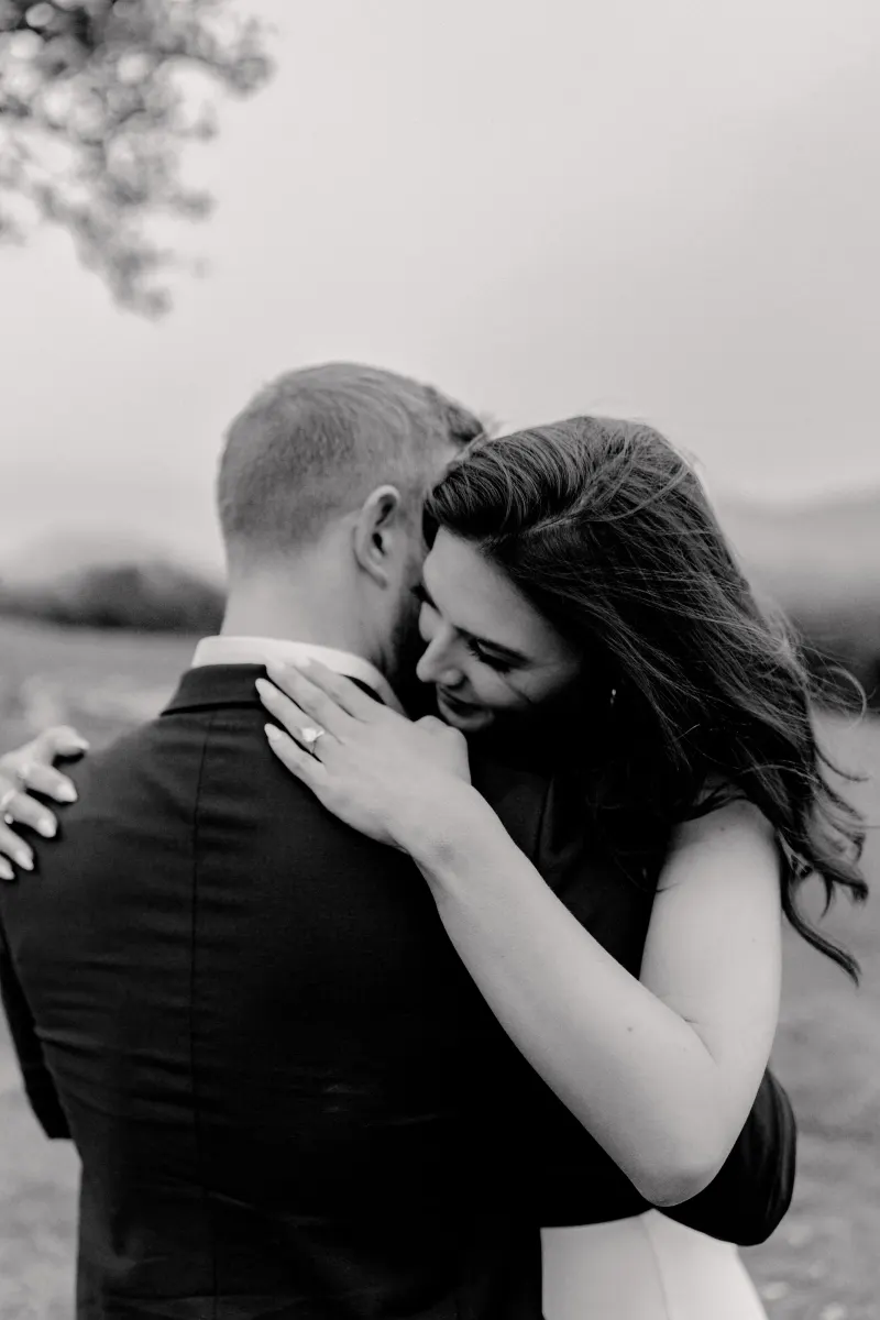 Bride-to-be smiles and hugs her fiancé, showing her engagement ring after a countryside surprise proposal photoshoot.