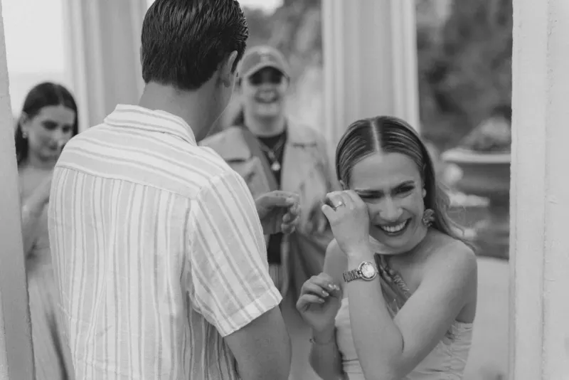 Bride-to-be wipes away happy tears while friends celebrate during a candid surprise proposal photoshoot by the sea.