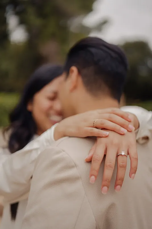 Bride-to-be wraps her arms around her fiancé, showing her engagement ring during a joyful couples photoshoot outdoors.