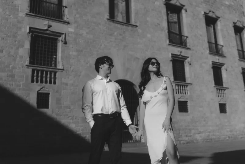 Chic couple holding hands and smiling in sunlight during a couples & engagements photoshoot by a historic stone building.