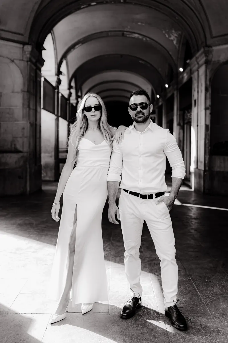 Chic couple in matching white outfits pose confidently under city arches during a couples & engagements photoshoot.