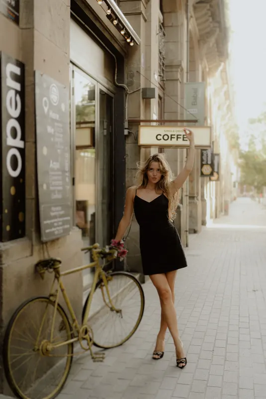 Chic woman in a black dress poses confidently by a café and vintage bike during a Vacation Portraits shoot in Europe.