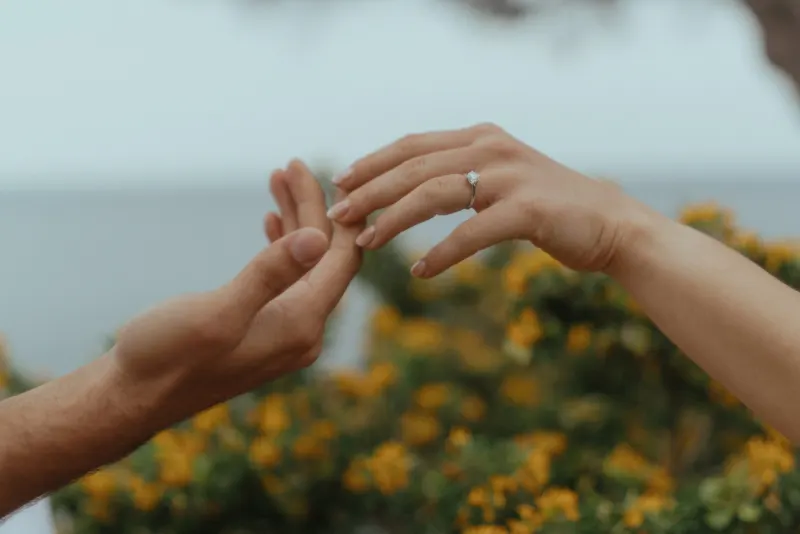 Close-up of a couple holding hands, engagement ring visible, during a seaside surprise proposal photoshoot.