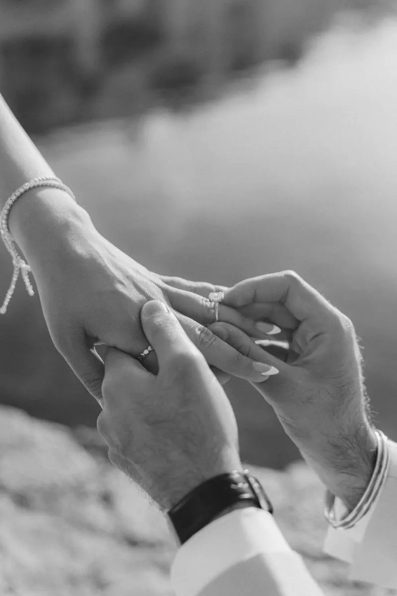 Close-up of a man placing an engagement ring on his partner’s hand during a surprise proposal photoshoot by the water.