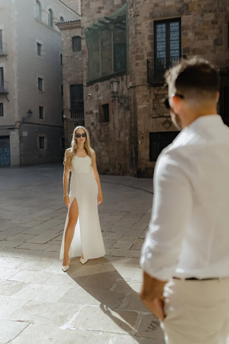 Confident couple in white share a sunlit moment in a historic city square during a couples & engagements photoshoot.