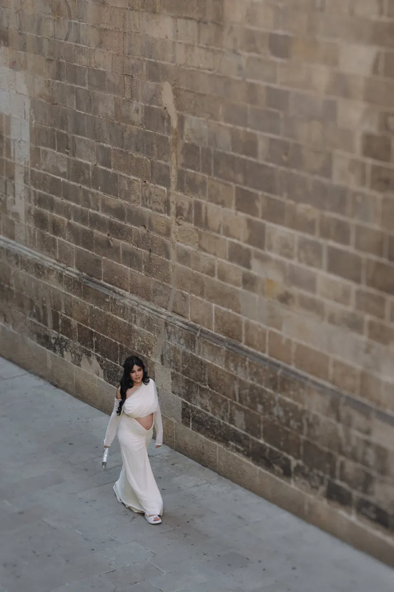 Confident expectant mother in a flowing white dress strolls beside a historic stone wall during a Vacation Portraits session.