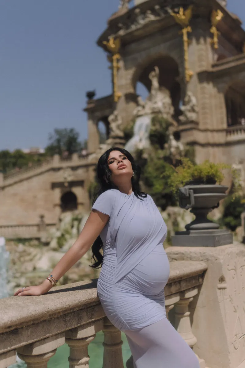 Confident pregnant woman in a pale blue dress leans on a stone railing at Parc de la Ciutadella, Vacation Portraits session.