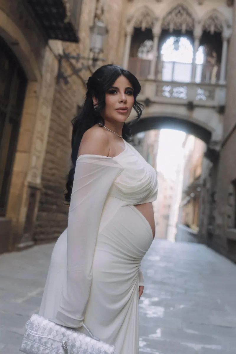 Confident pregnant woman in a white dress poses in a charming old city street during a Vacation Portraits session.