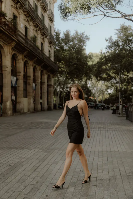 Confident woman in a black dress walks down a sunlit European street during a Vacation Portraits photoshoot.