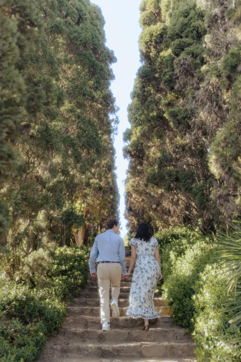Couple ascends sun-dappled garden steps hand in hand, captured during a surprise proposal photoshoot.
