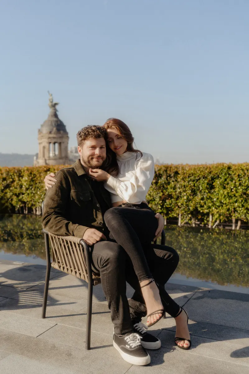 Couple cuddles on a rooftop chair, sharing a tender moment during an engagement session with city views.