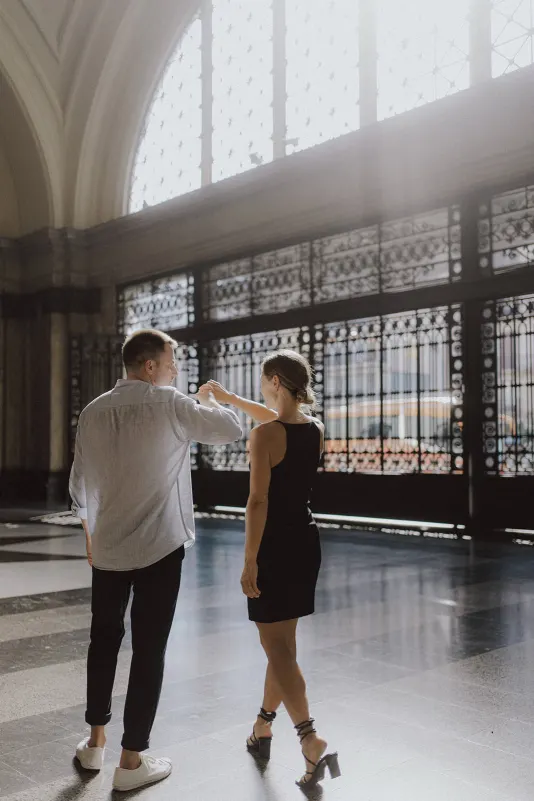 Couple dances together in a sunlit grand hall, sharing a playful moment during a couples & engagements photoshoot.