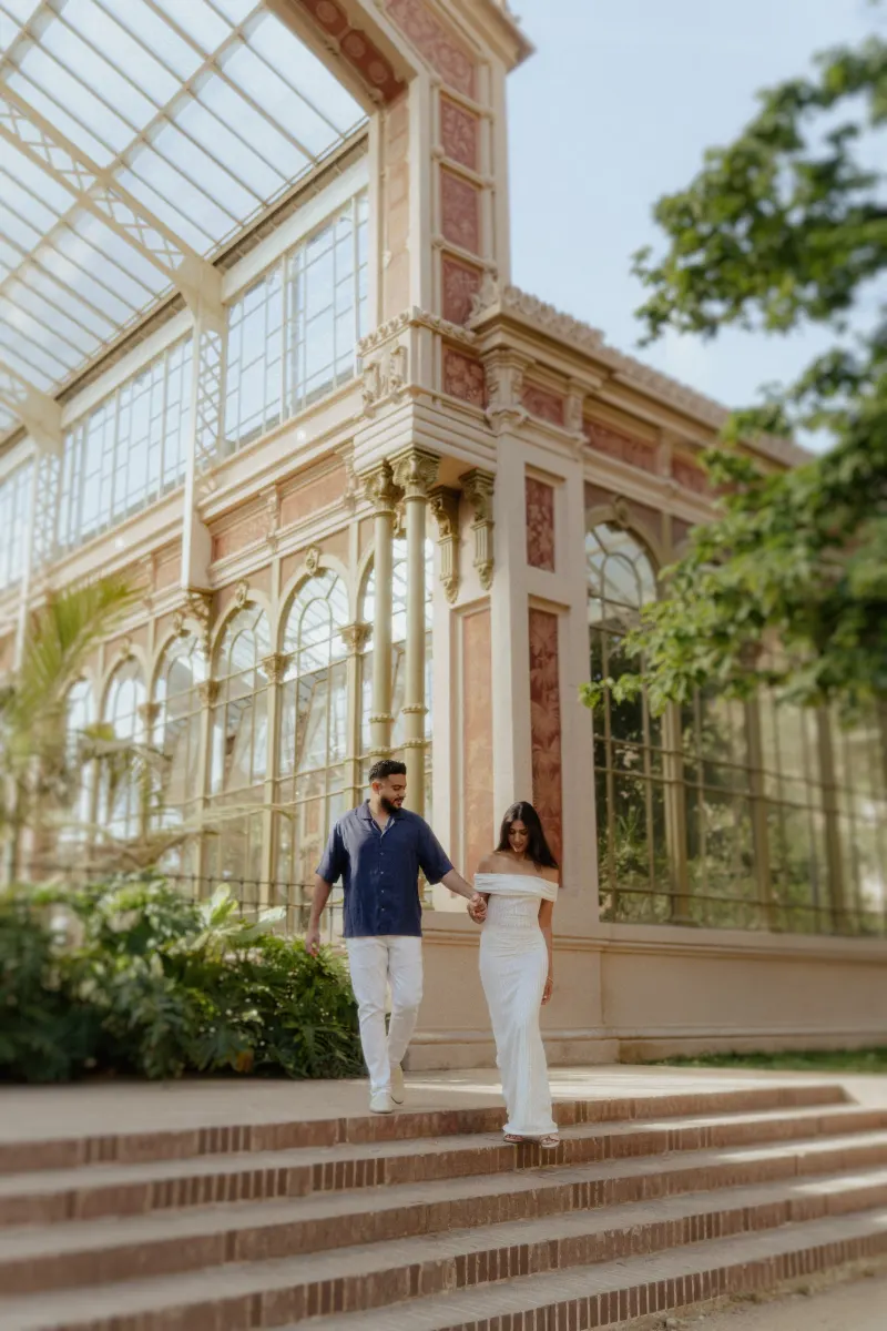 Couple descending garden steps hand in hand, sharing a tender moment during an engagement shoot at a grand glass conservatory