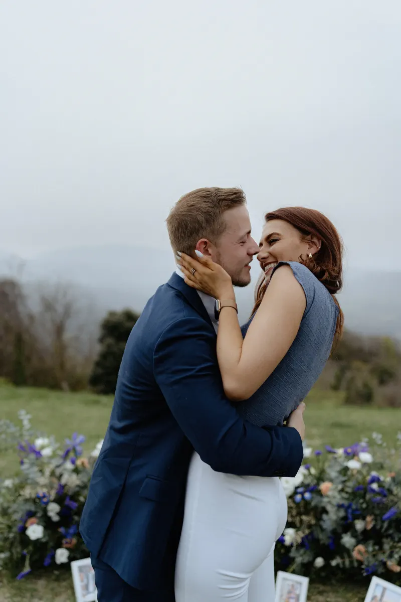 Couple embraces and laughs after a surprise proposal on a flower-filled hillside, captured during a proposal photoshoot.