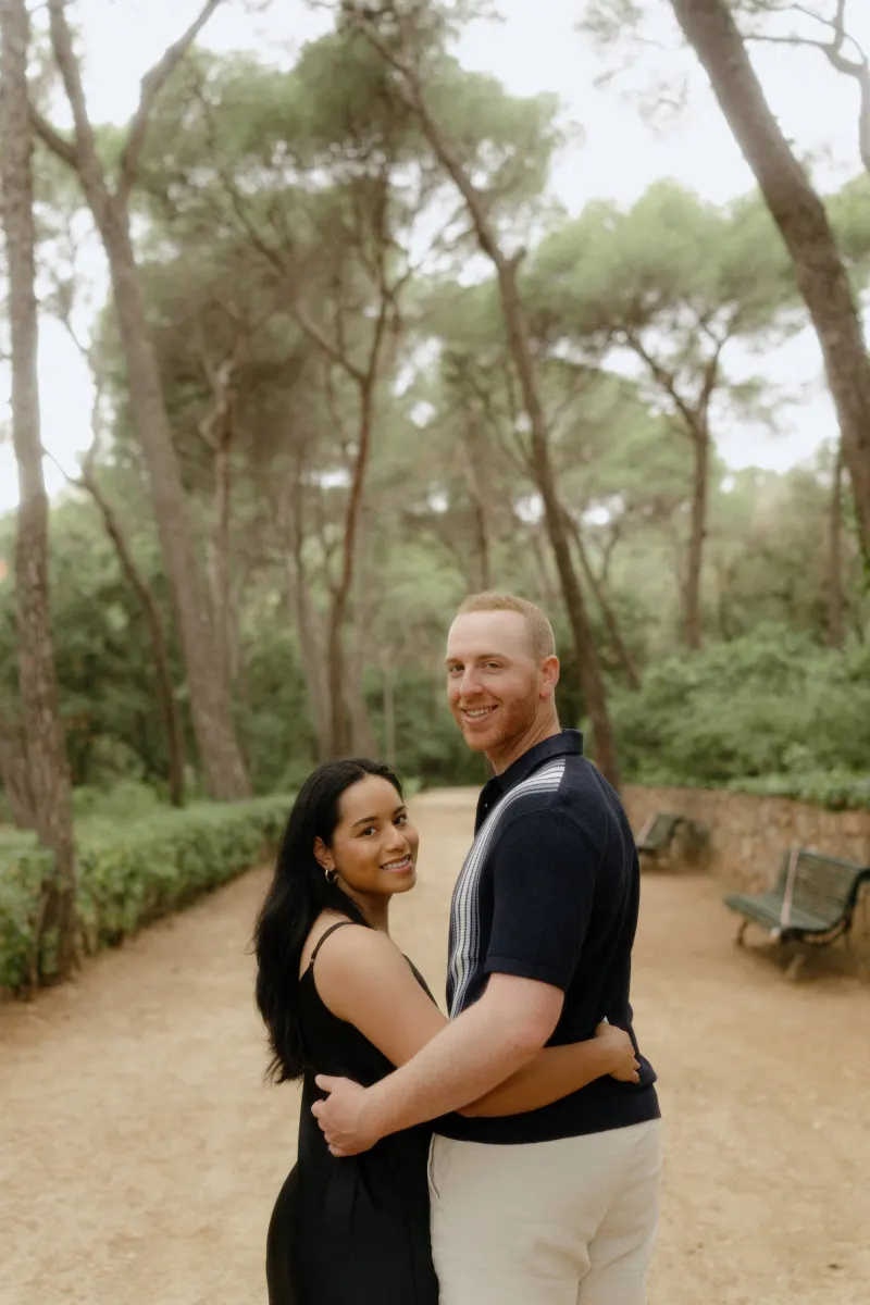 Couple embraces and smiles at the camera on a tree-lined path during a surprise proposal photoshoot in the park.