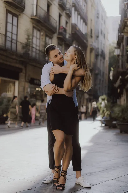 Couple embraces and smiles on a sunlit city street, sharing a joyful moment during a couples & engagements photoshoot.