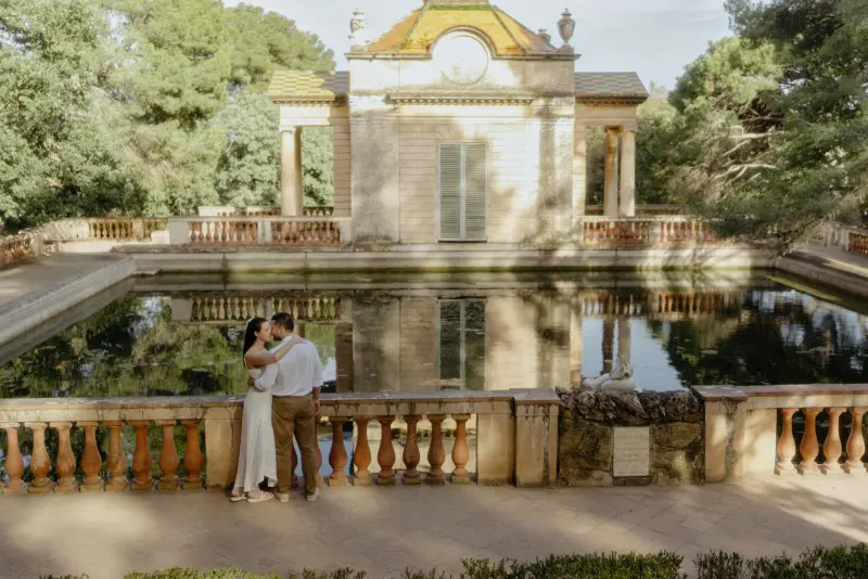 Couple embraces by a tranquil pond in front of a classical pavilion during a romantic couples & engagements photoshoot.