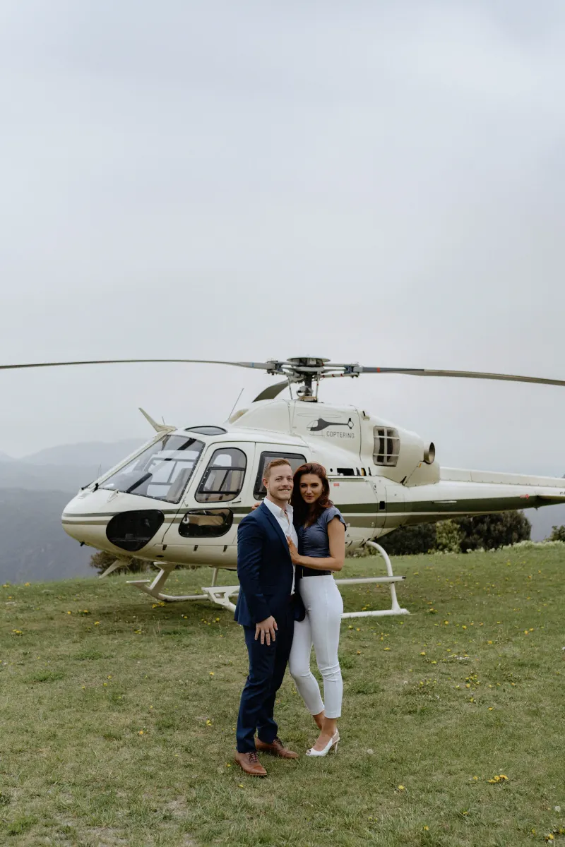 Couple embraces on a grassy hillside in front of a helicopter, celebrating their countryside surprise proposal photoshoot.