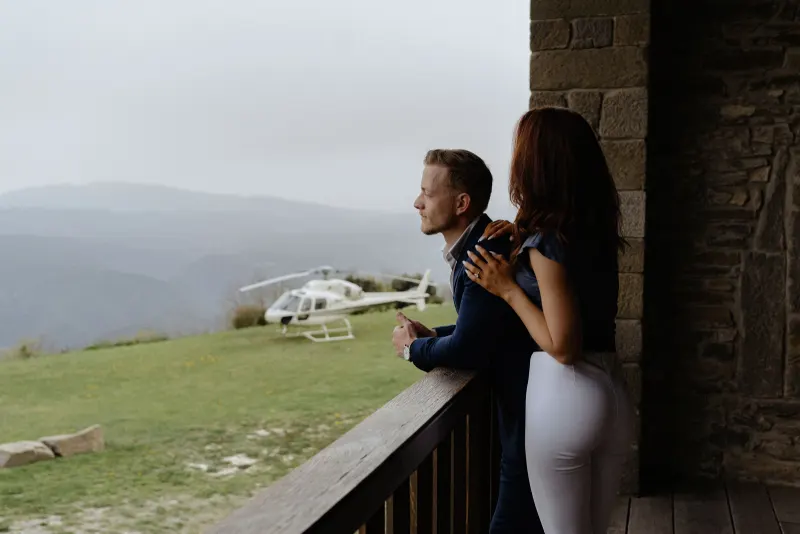 Couple embraces on a rustic balcony, gazing at mountain views and a helicopter after their surprise proposal photoshoot.