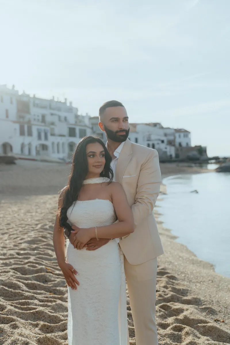 Couple embraces on a sunlit beach during a surprise proposal photoshoot, with white coastal buildings in the background.