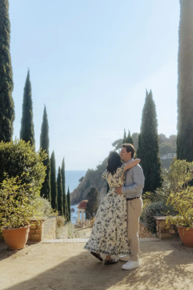 Couple embraces on a sunlit garden path overlooking the sea during a surprise proposal photoshoot.