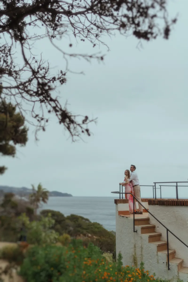 Couple embraces on a terrace overlooking the ocean, sharing a joyful moment during a surprise proposal photoshoot.