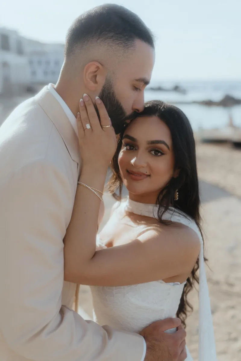 Couple embraces on the beach after a surprise proposal, bride-to-be smiling and showing her engagement ring.
