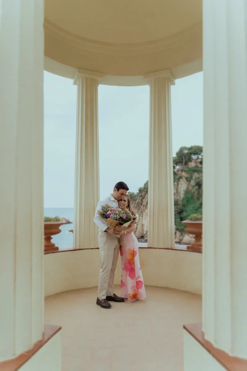 Couple embraces with a bouquet in a seaside pavilion, celebrating their engagement during a surprise proposal photoshoot.