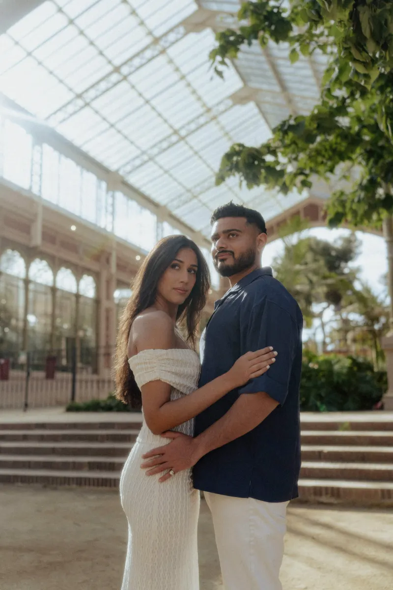 Couple embracing and gazing softly at the camera during an engagement photoshoot in a sunlit glass conservatory.