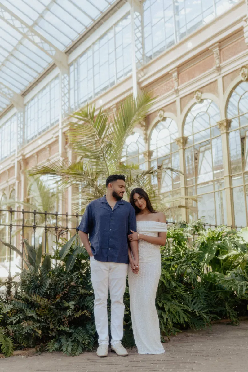 Couple embracing gently among lush greenery during an engagement photoshoot in a sunlit glass conservatory.