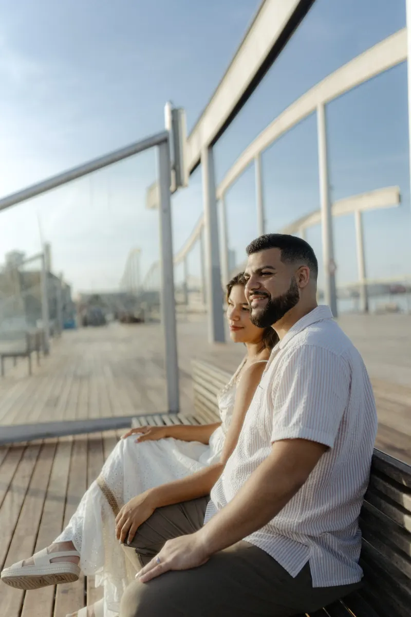 Couple enjoys a relaxed moment together on a sunlit boardwalk bench during their engagement photoshoot by the marina.