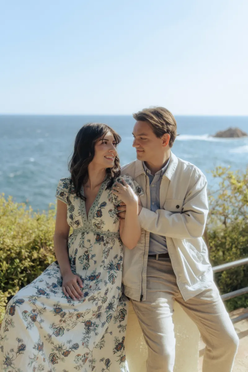 Couple gazes at each other, sharing a joyful moment by the ocean during a surprise proposal photoshoot.